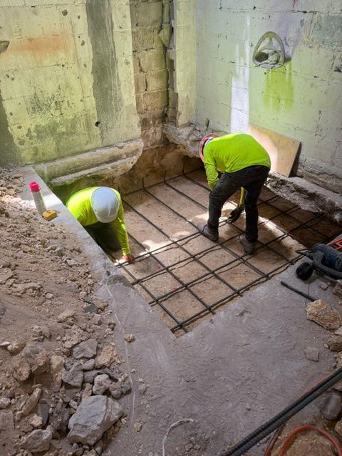 Two workers in neon high-visibility shirts install a metal rebar grid on a dirt floor inside a building undergoing repair.