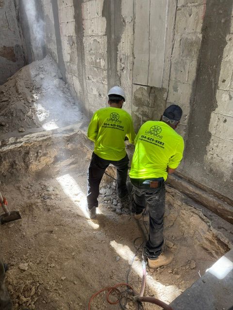 Two workers in bright neon yellow high-visibility shirts use jackhammers to break up concrete in an unfinished room.