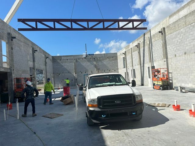 A crane lifts a steel truss over a construction site with workers near a white pickup truck.