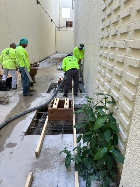 Four workers in lime-green shirts work in a narrow alley on a concrete construction project involving a wooden frame.