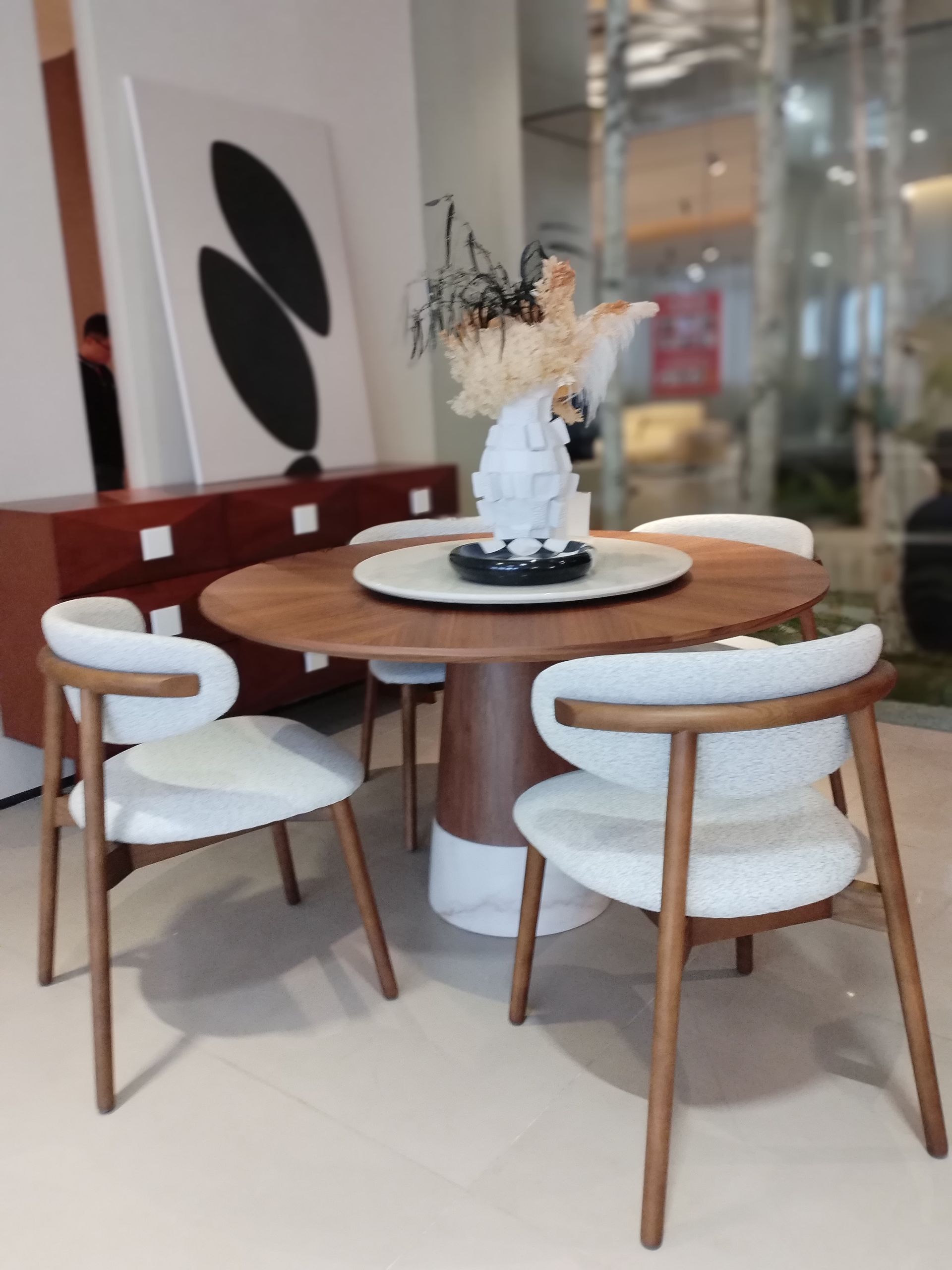 Dining room set: round wooden table, white upholstered chairs, floral arrangement, and abstract artwork.