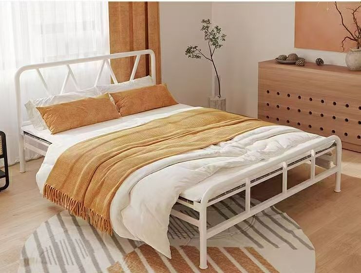 White metal bed frame with orange bedding, in a bright room.
