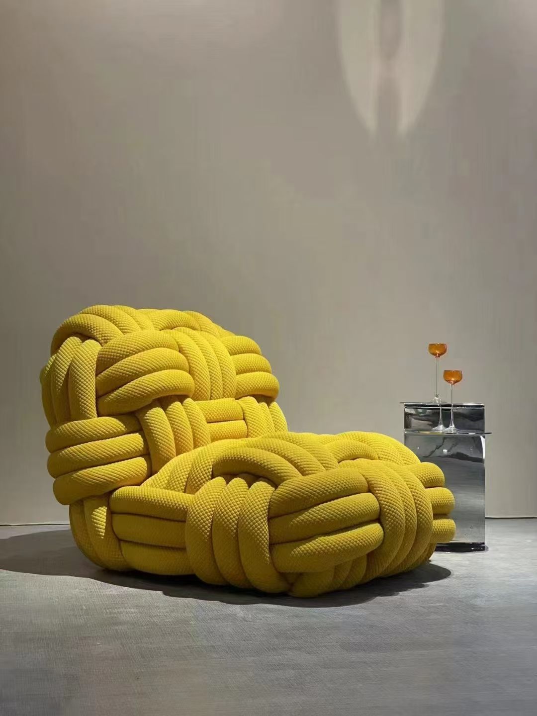 Yellow knotted fabric armchair next to a silver cube table, against a plain white wall.
