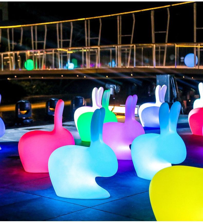 Colorful, glowing, rabbit-shaped chairs arranged outdoors at night. Lit walkway and glowing orbs in the background.