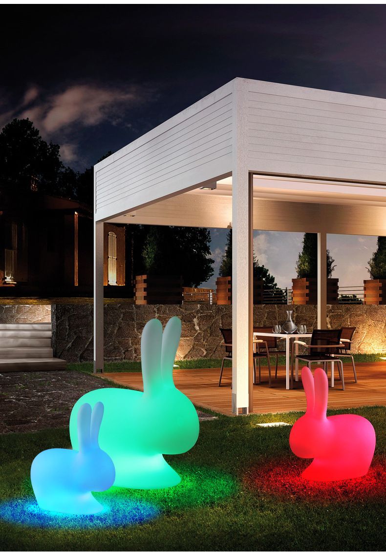 Three illuminated rabbit sculptures in blue, green, and pink on a lawn, near a white pergola at night.