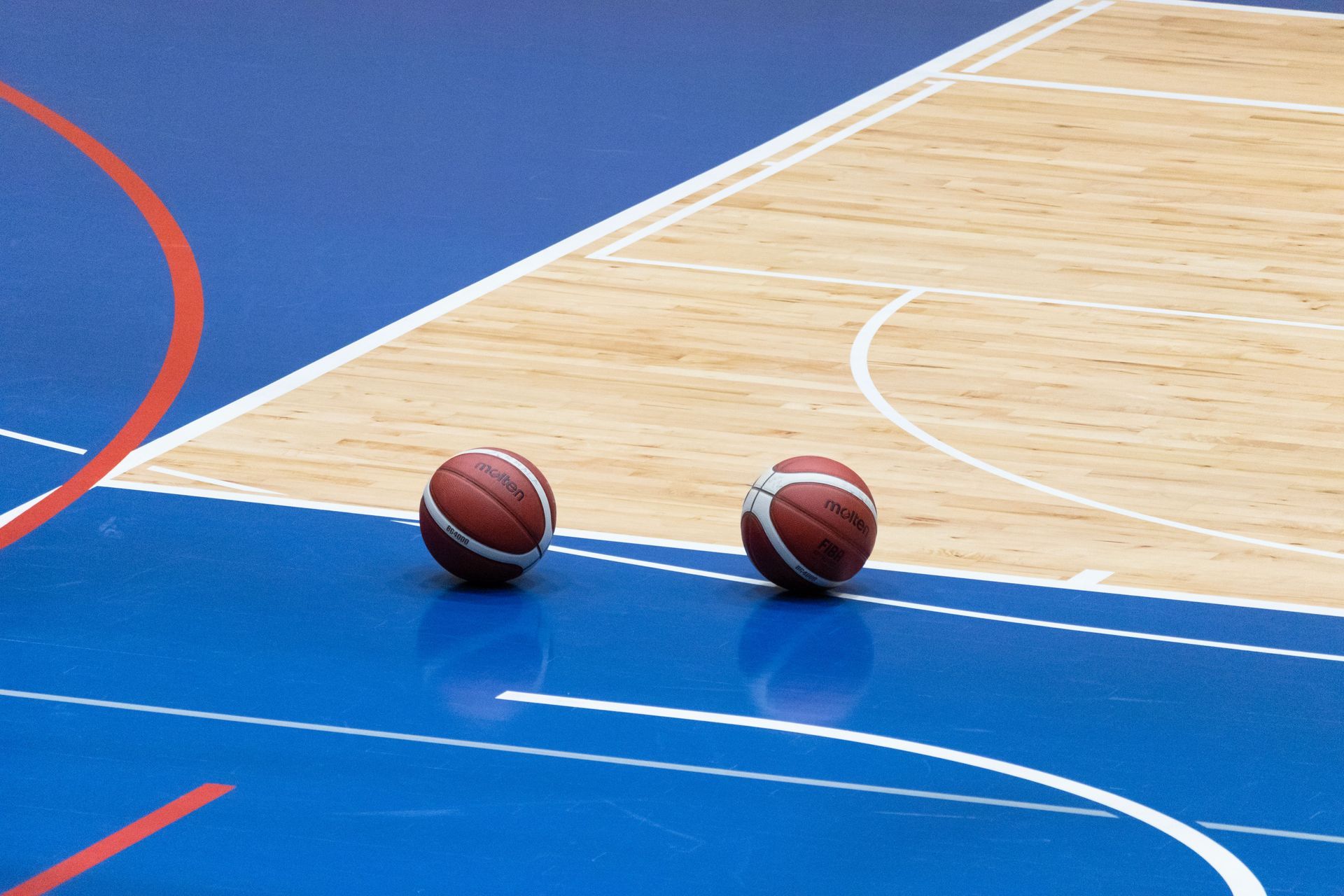 Two basketballs on a blue indoor court near the sideline, with white and red markings