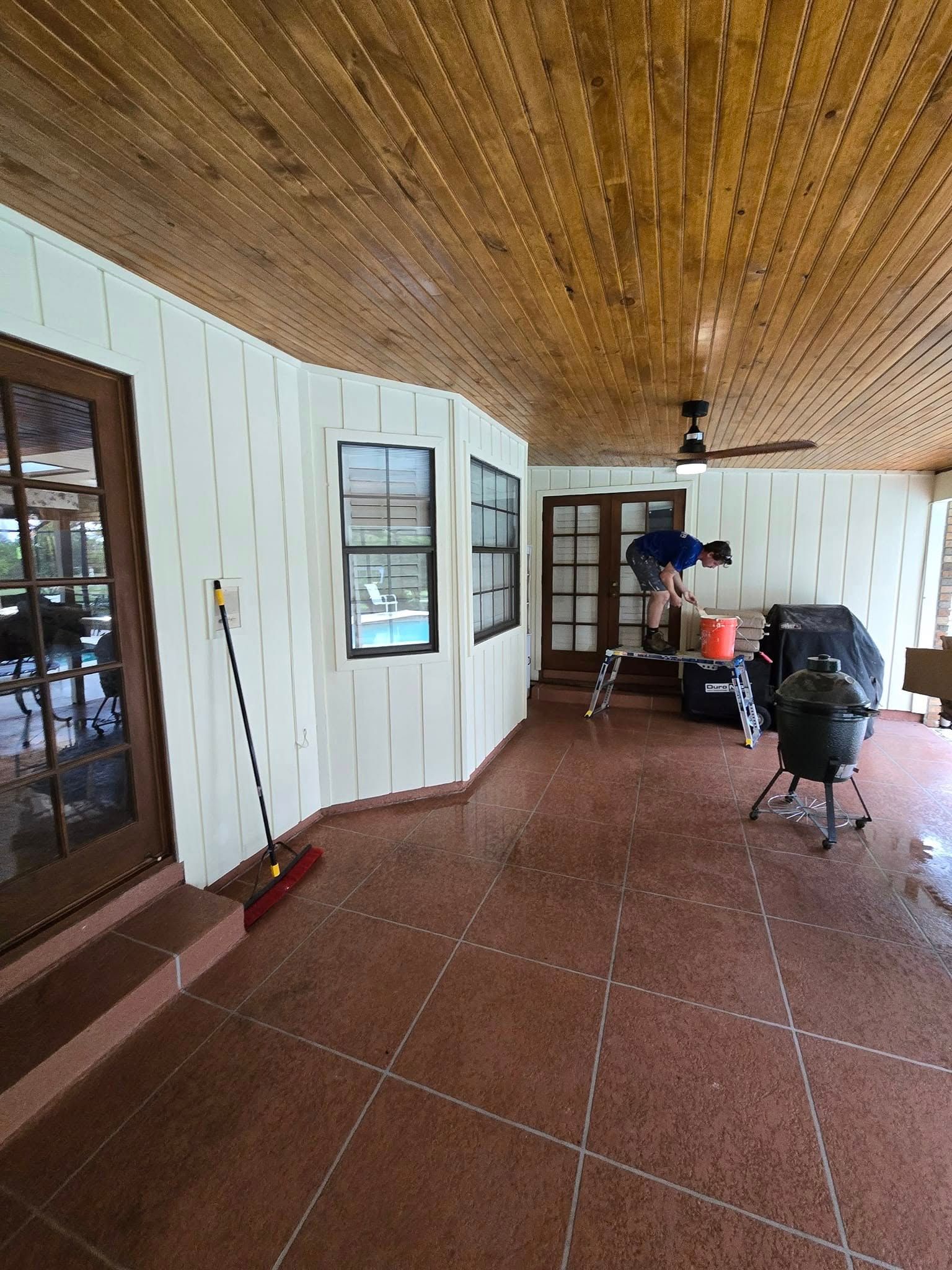 Covered patio with wood ceiling, red tile floor, white walls, two windows, and a black grill.