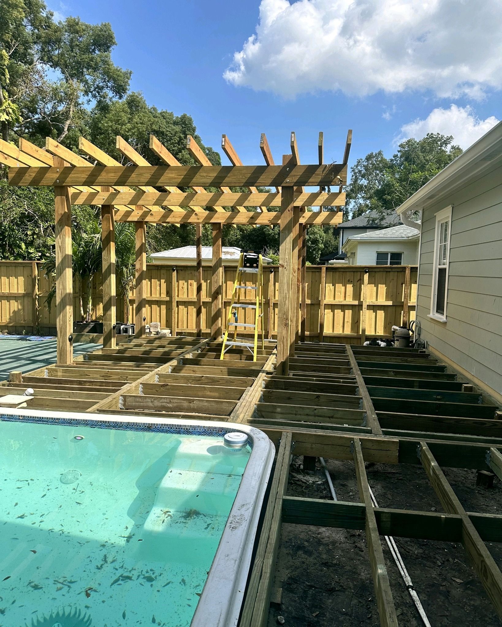 Pergola construction over a deck next to a pool. Wooden fence and house in the background under a blue sky.