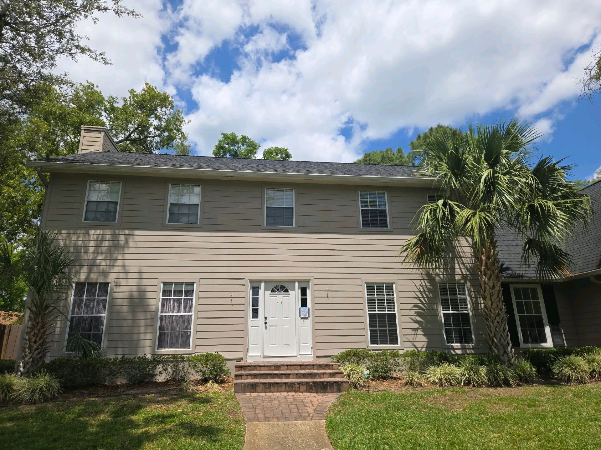 Two-story beige house with a white front door and several windows, palm tree on the right, under a blue sky.