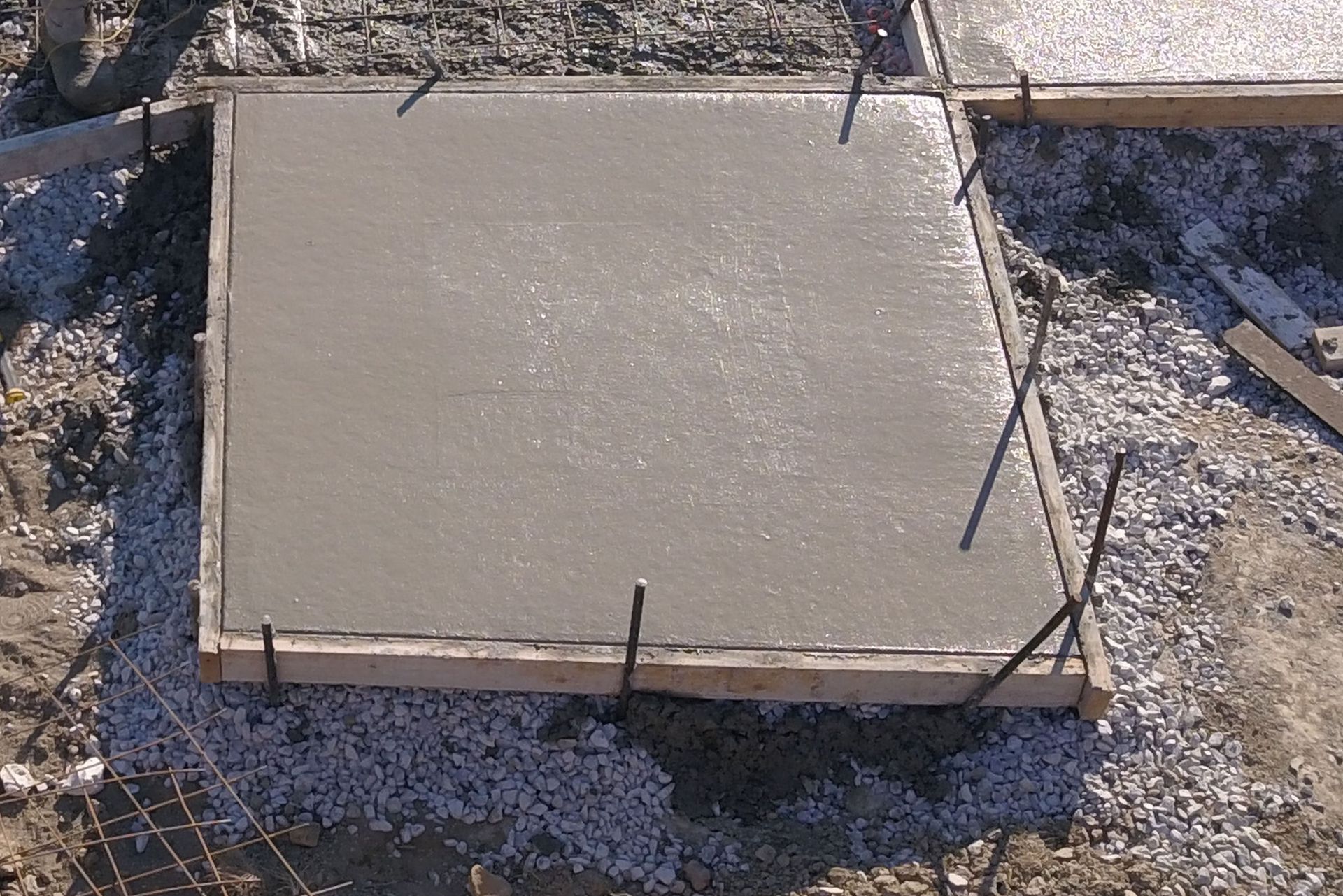 Freshly poured concrete slab within a wooden form, surrounded by gravel and rebar.