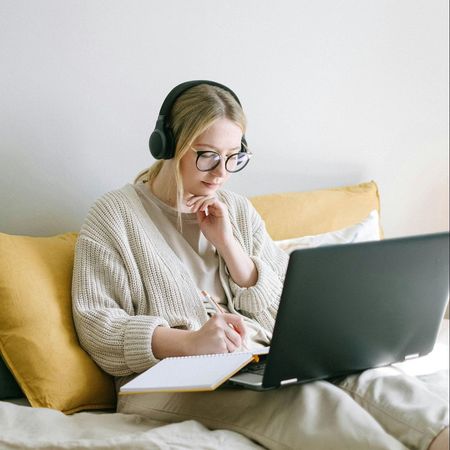 A woman wearing headphones is sitting on a bed using a laptop computer.