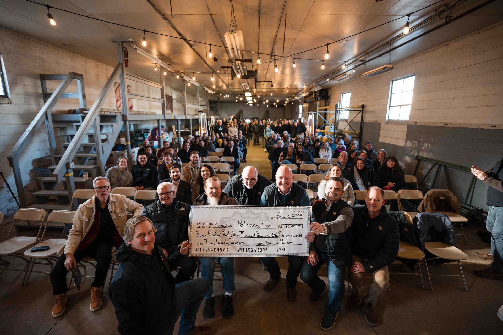 Large group of people in an open space, holding a sign. Men in foreground smiling, others in rows of chairs.