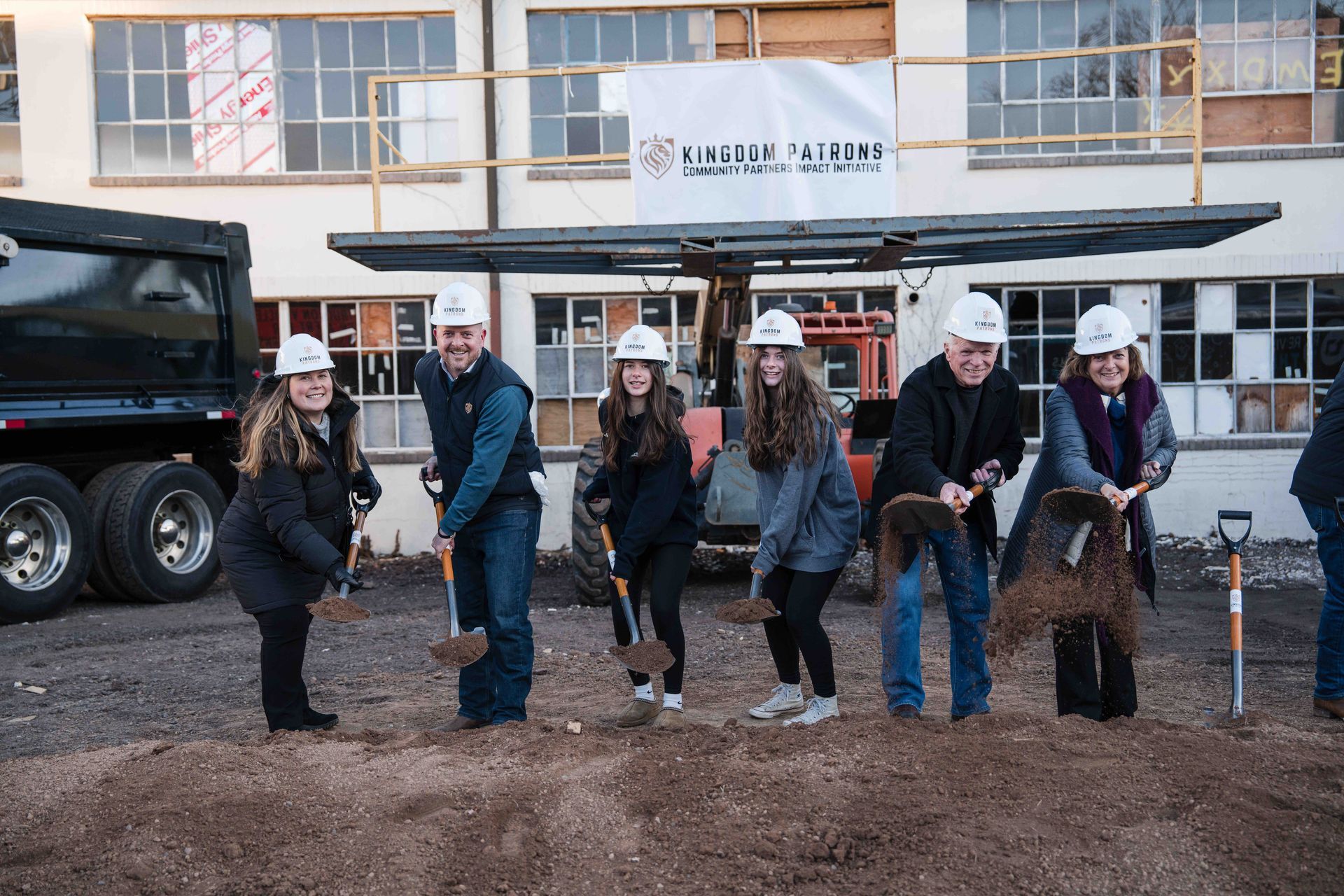 People in hard hats and casual attire at a groundbreaking ceremony, digging with shovels. Building in background.