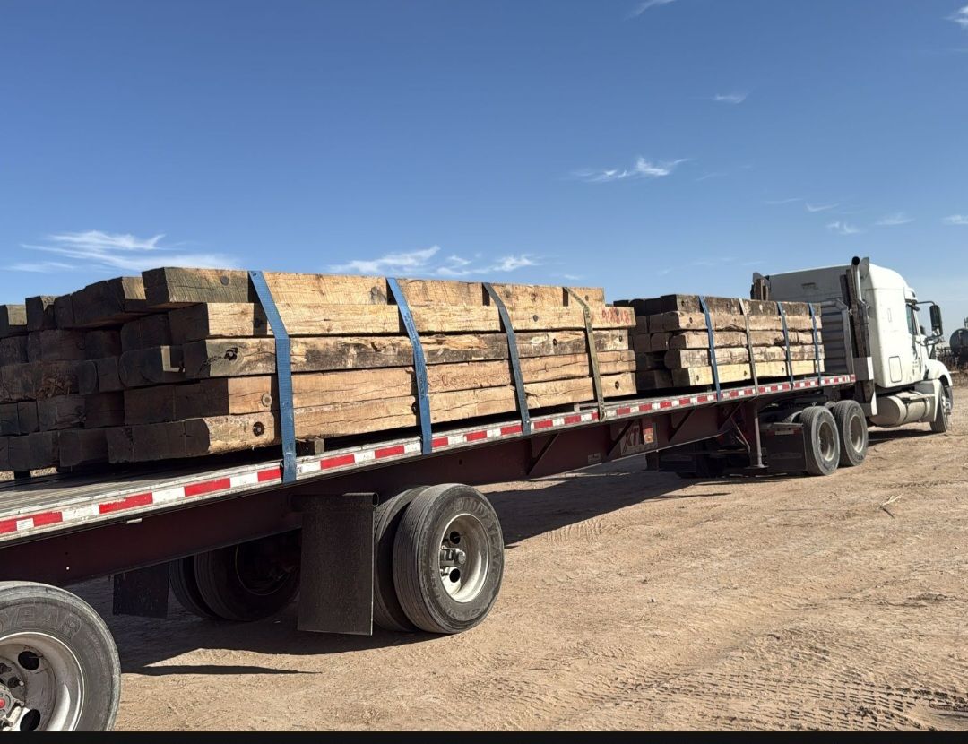 Flatbed truck loaded with strapped crane mats on a dirt lot under a clear blue sky