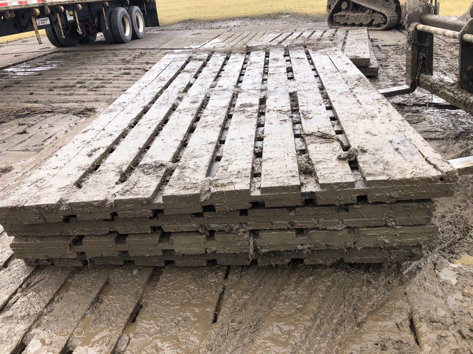 Stacked muddy ground protection mats on a construction site, with a truck and excavator in the background.