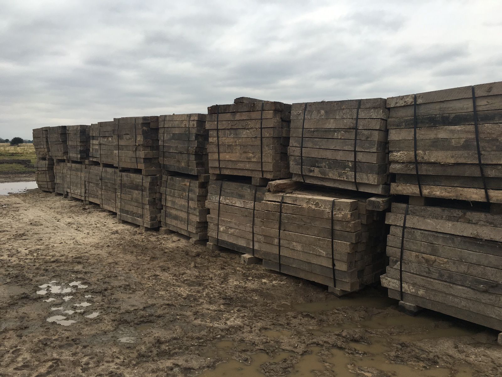 Stacks of used pipeline skids for sale on muddy ground under a cloudy sky.