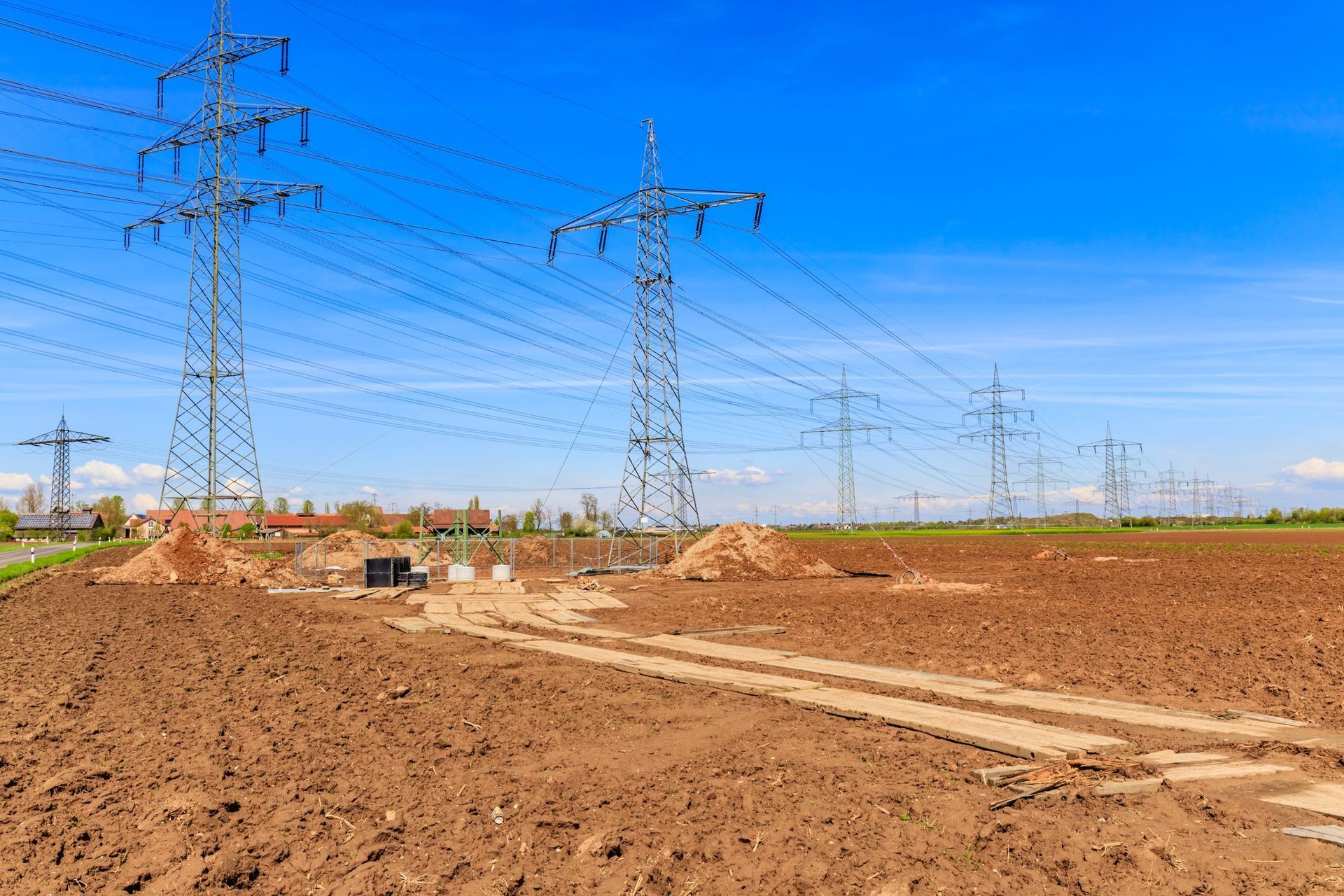 Electric transmission towers over a freshly graded dirt field under a bright blue sky with mat access for power