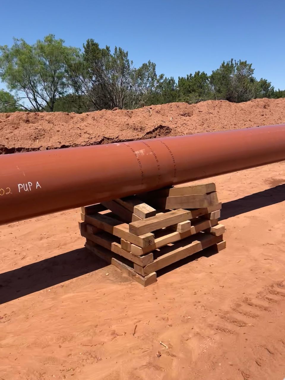 Large rust-colored pipe resting on pipeline skids in a red dirt construction area outdoors.