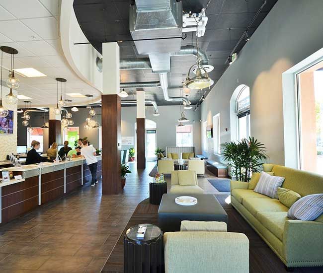 Reception area: Light green sofa, chairs, brown carpet, check-in counter, gray walls, dark ceiling, employees assisting clients.