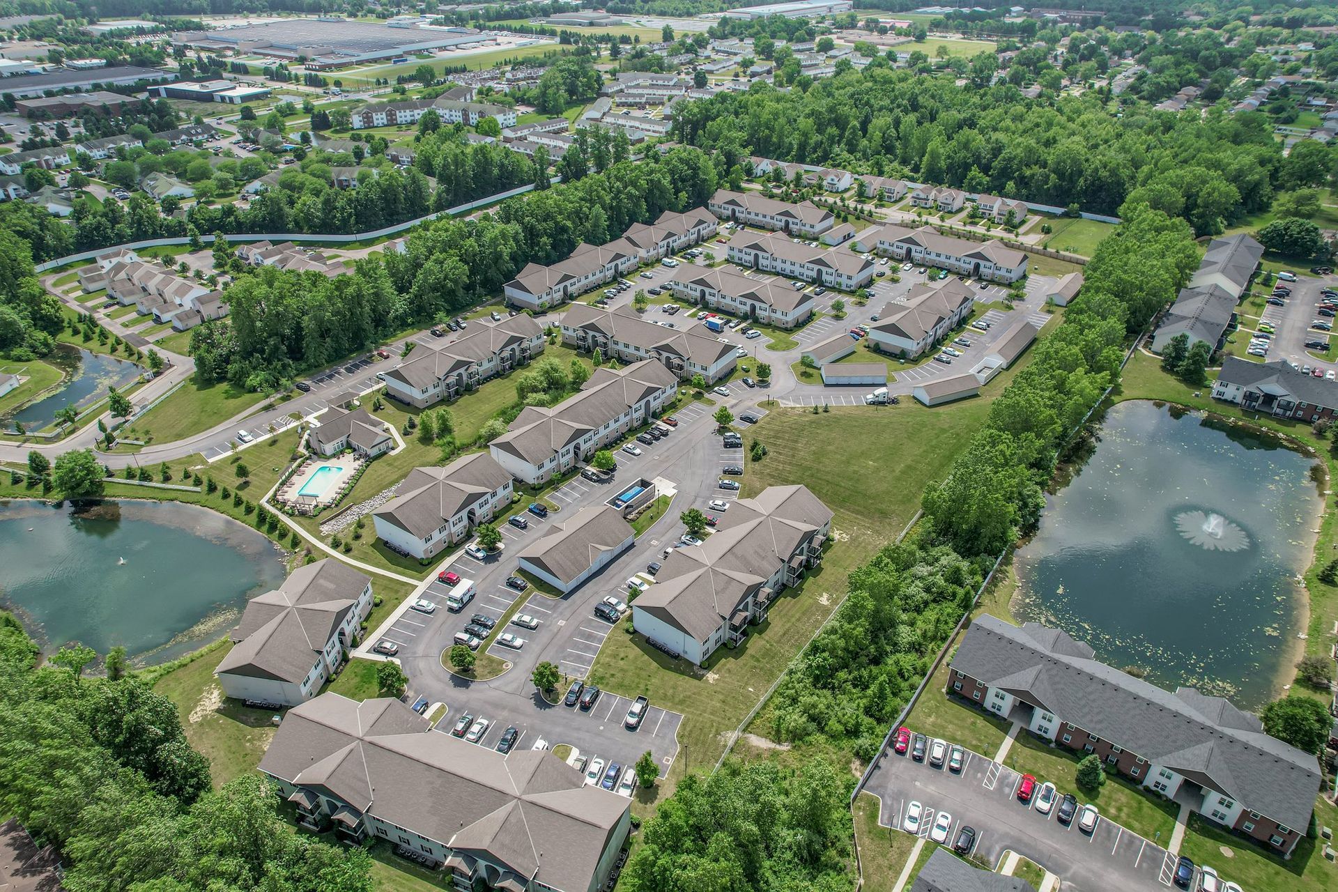 Aerial view of a suburban apartment complex with parking lots, roads, and surrounding green trees and ponds