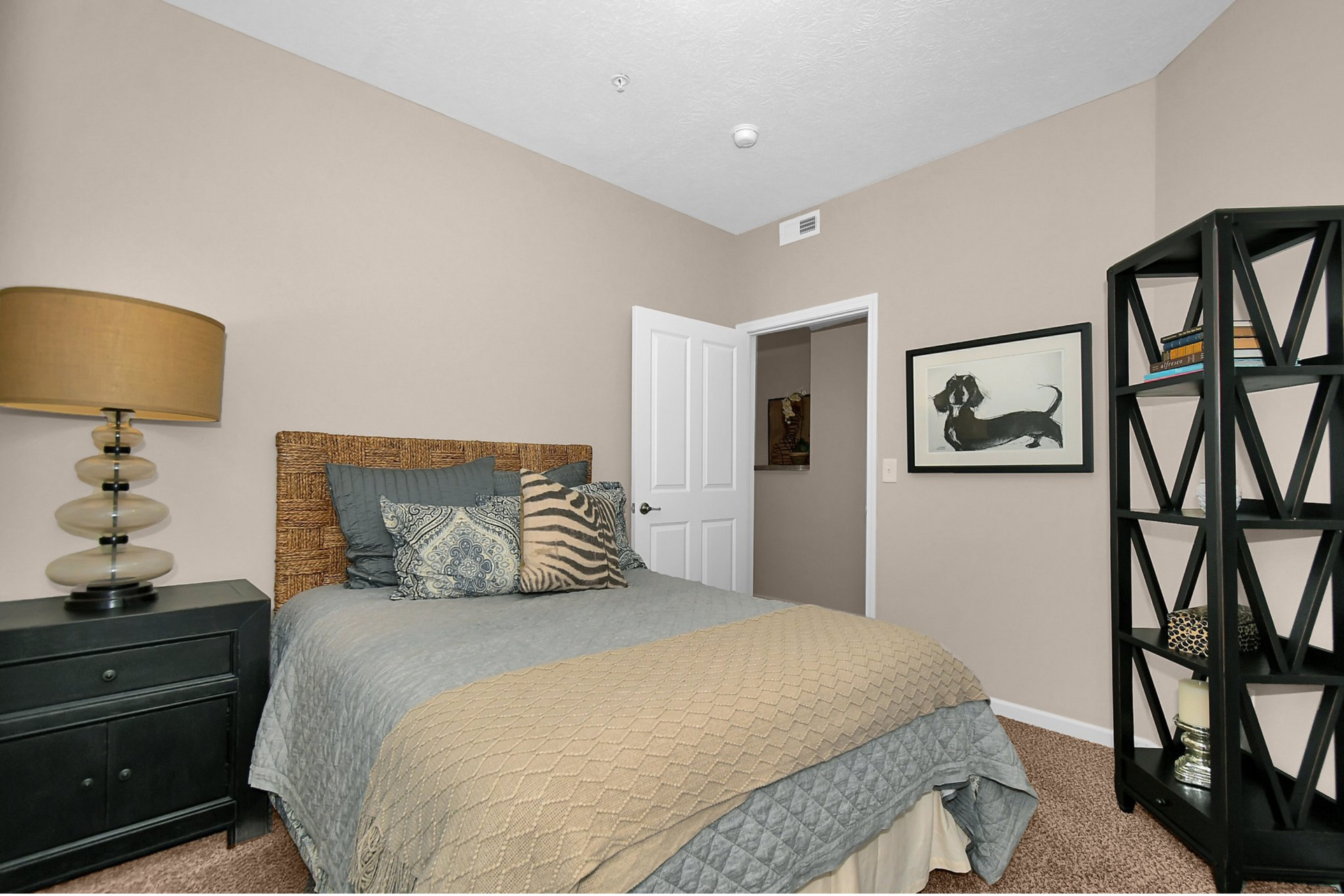 Neatly made beige bedroom with dark dresser, patterned headboard, wall art, and open white door