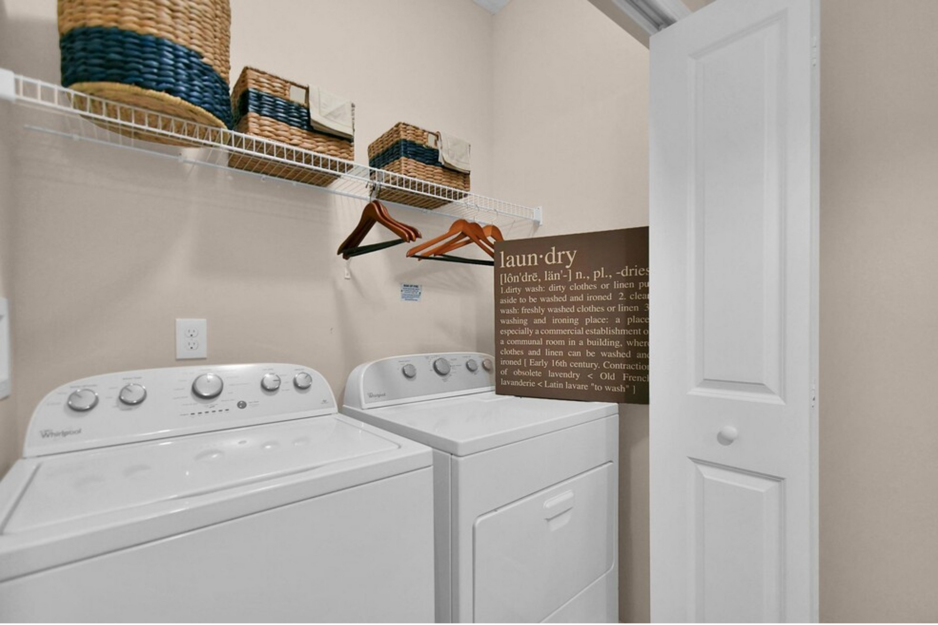 Laundry room with white washer and dryer, wall shelves, hanging rods, baskets, and a decorative sign.