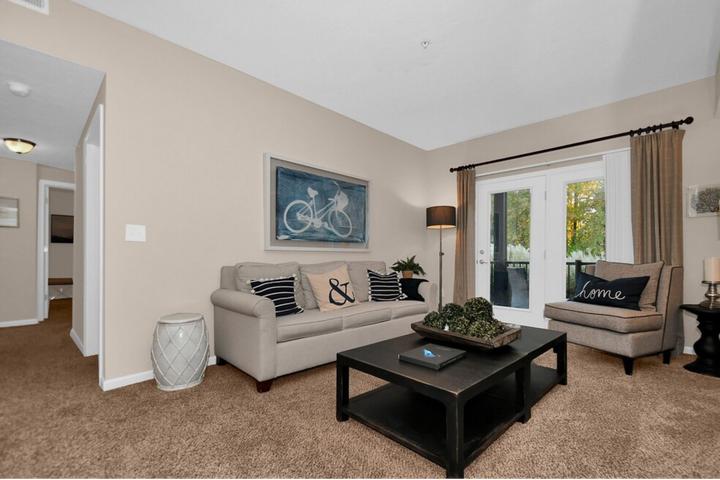 Bright living room with beige walls, sofa, coffee table, lamps, and a dining area in the background