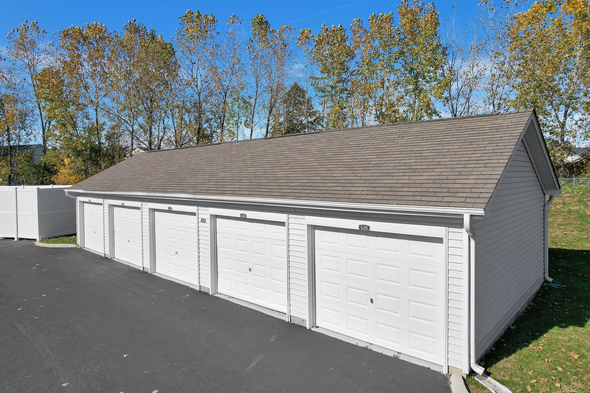 Row of white storage garage units with closed doors beside a paved lot and trees
