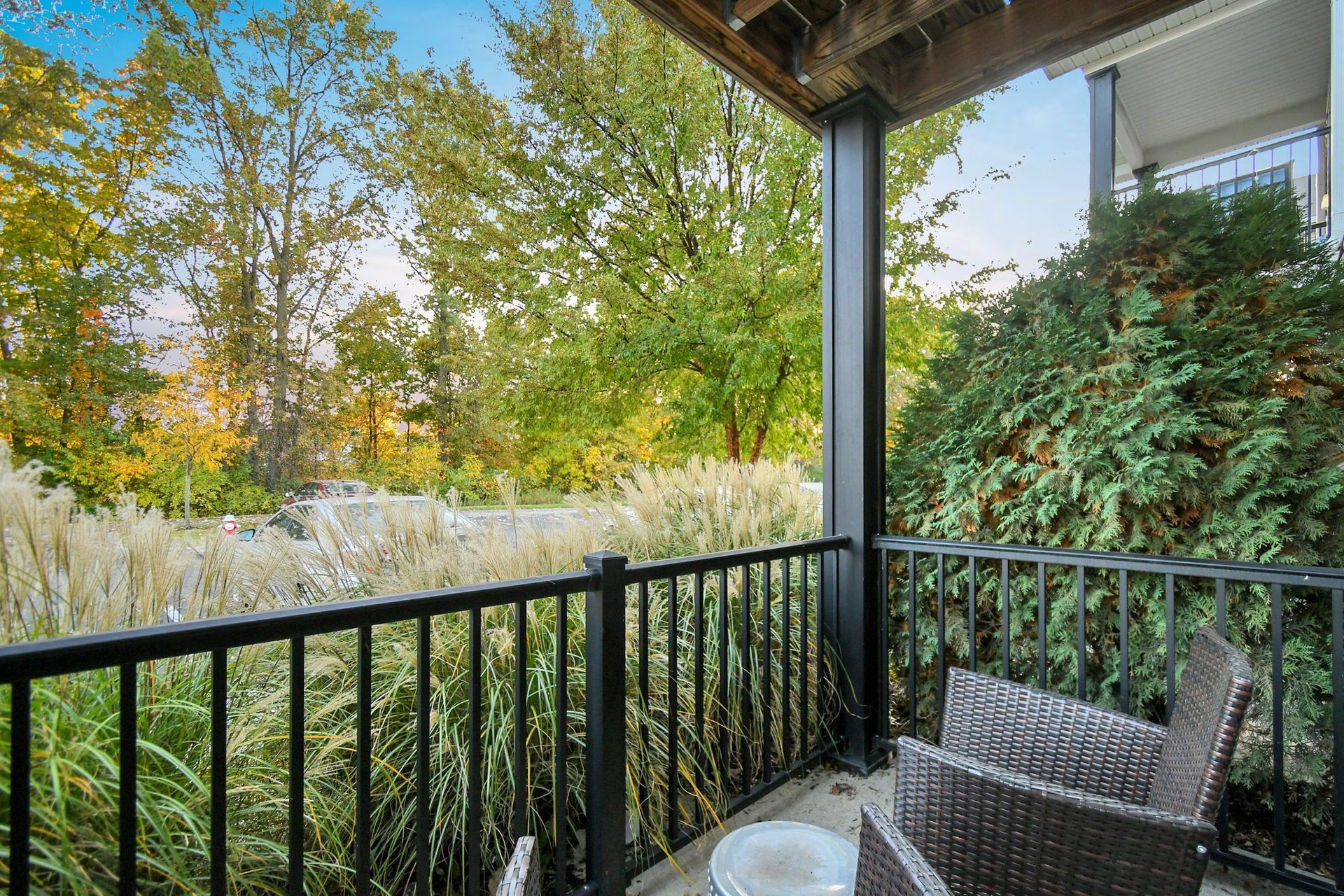 Covered balcony with black railing, wicker chair, and a view of green trees and a bright garden.