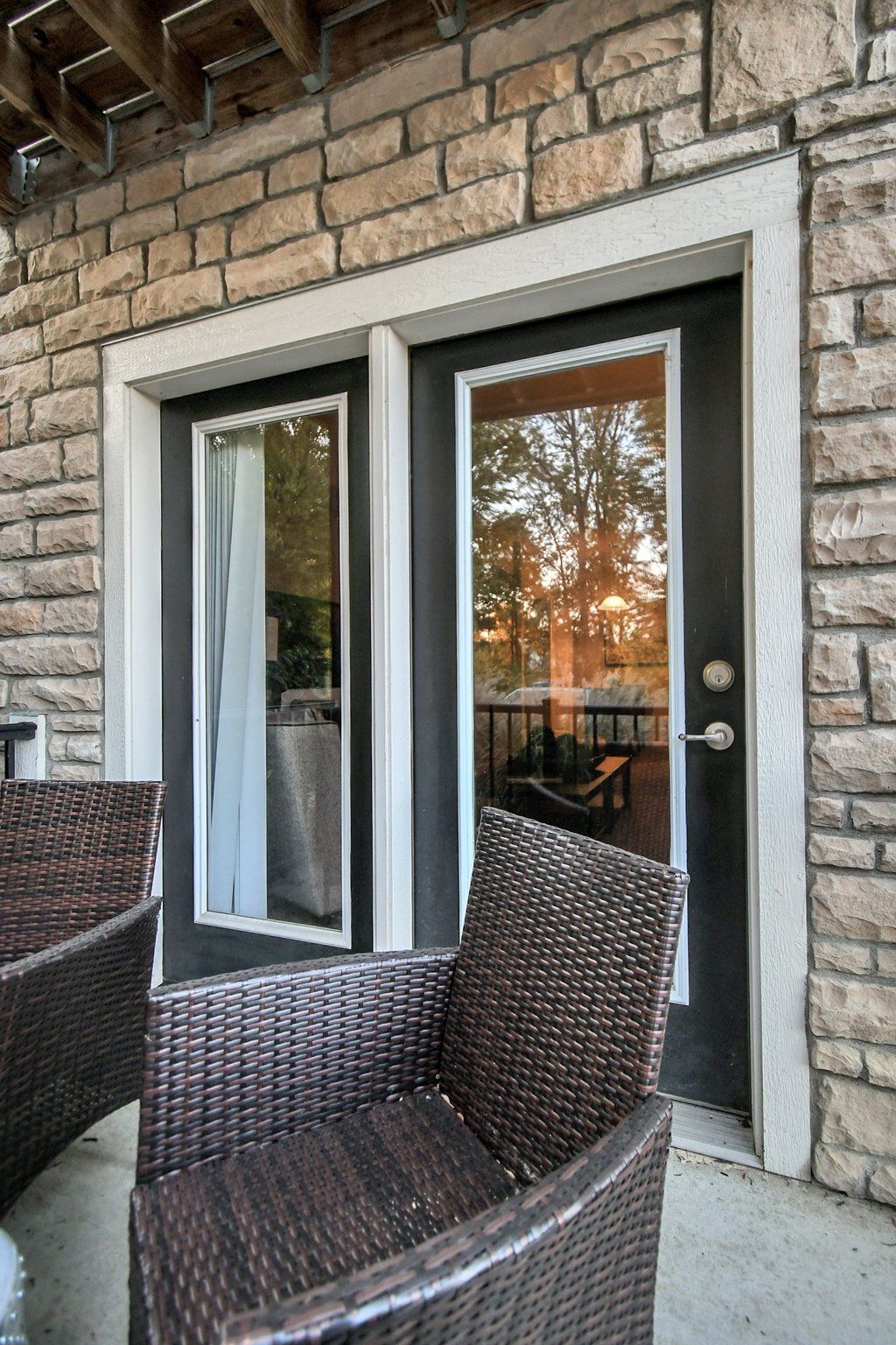 Wicker chairs on a stone patio beside a glass door and window under a brick exterior wall at sunset