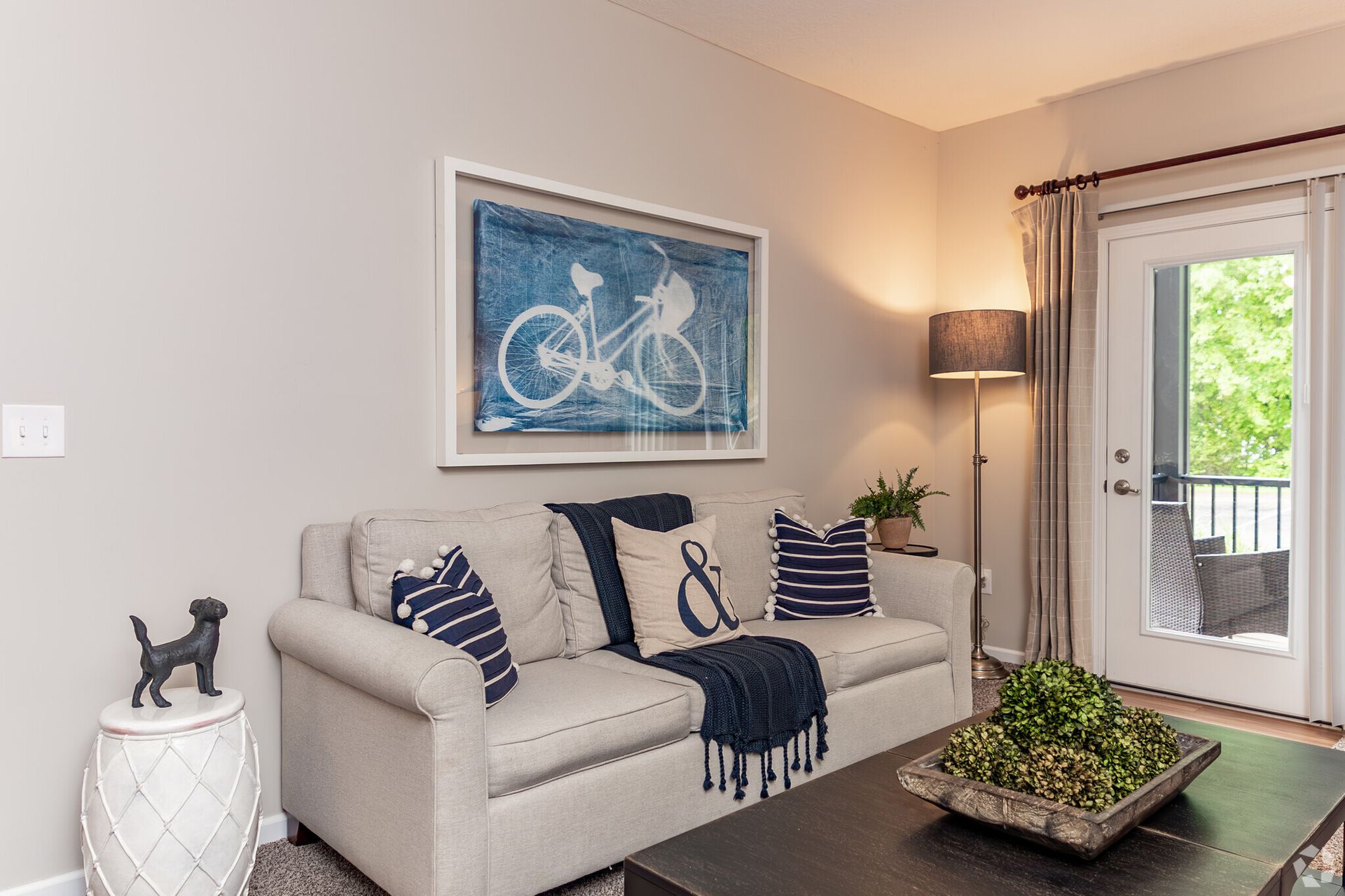 Cozy living room with beige sofa, blue bicycle wall art, floor lamp, and glass door to a patio with greenery