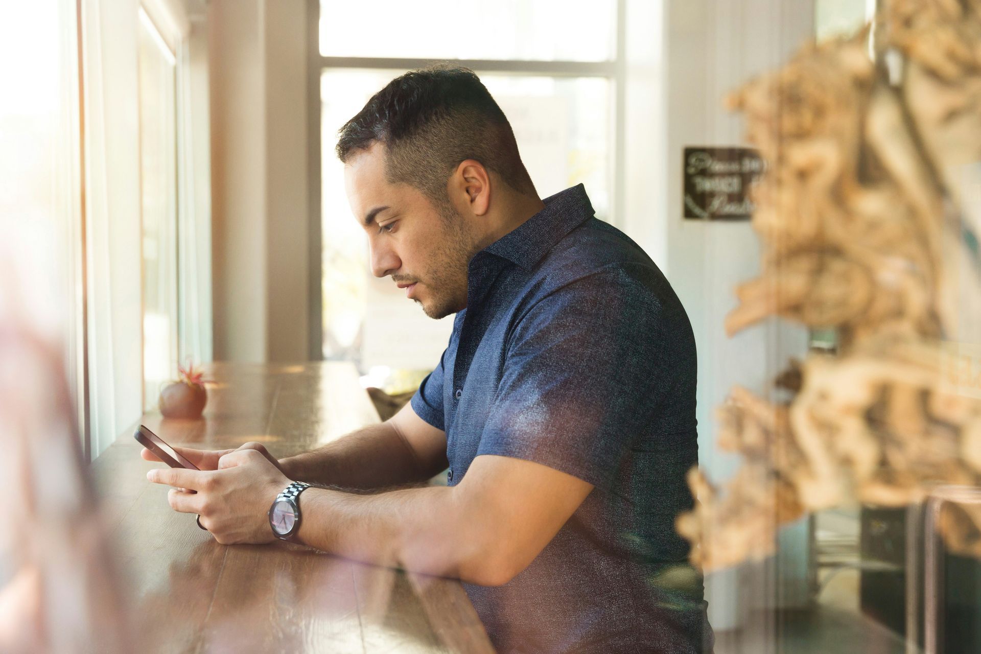 Man in a blue shirt looking at his phone while leaning by a sunlit window