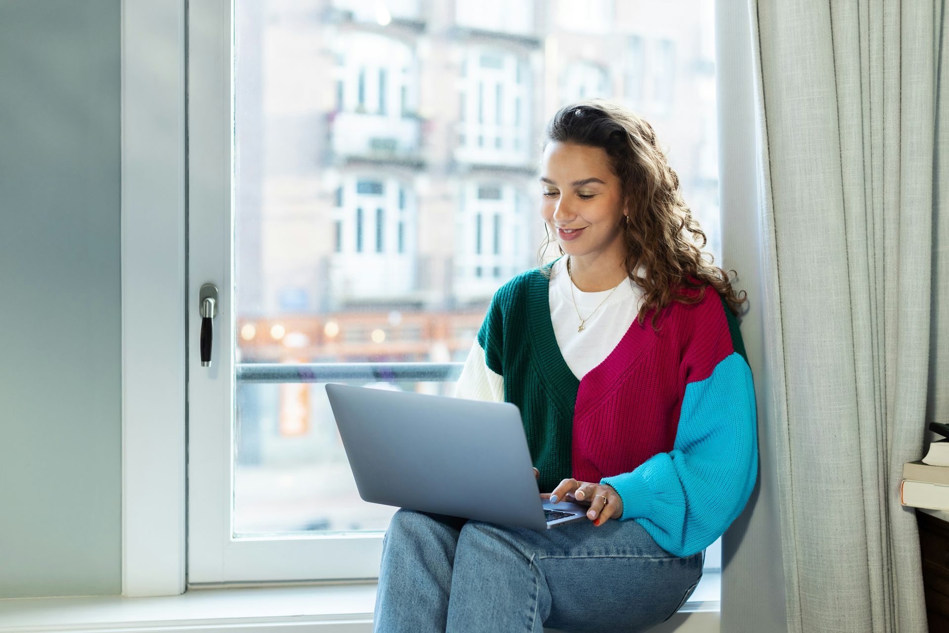 Woman using a laptop by a window, wearing a green, pink, and blue sweater.