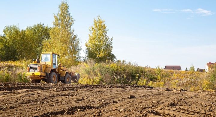 Yellow bulldozer grading a dirt field with trees and a building in the background
