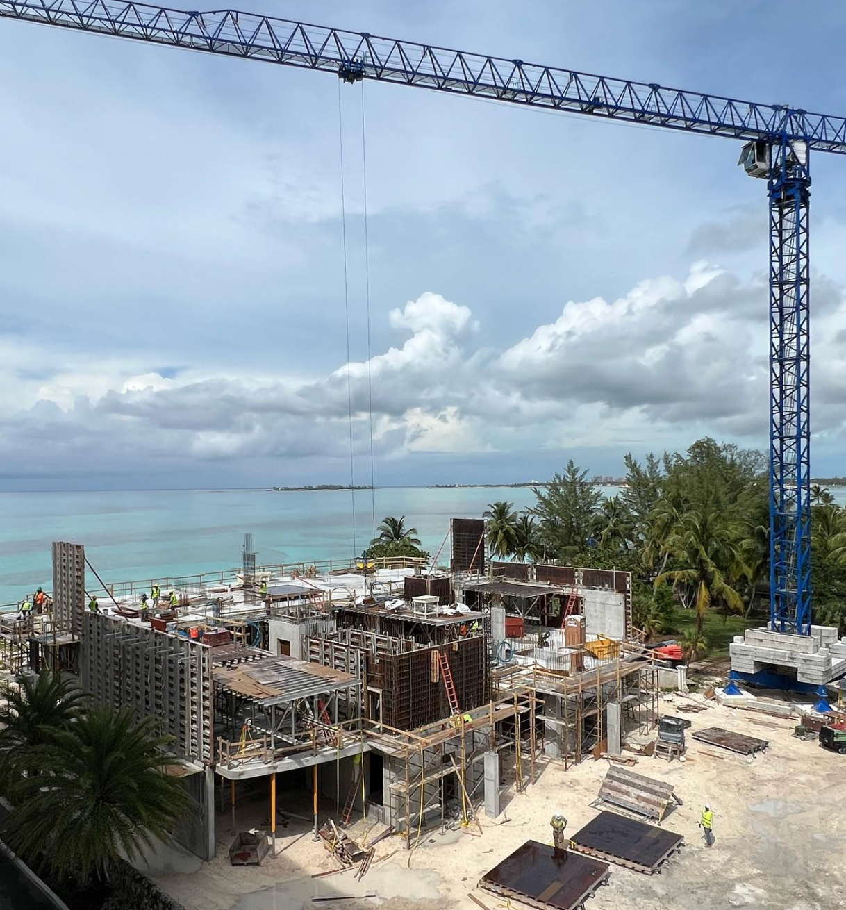 Construction site by the ocean, with crane, cloudy sky, and turquoise water.