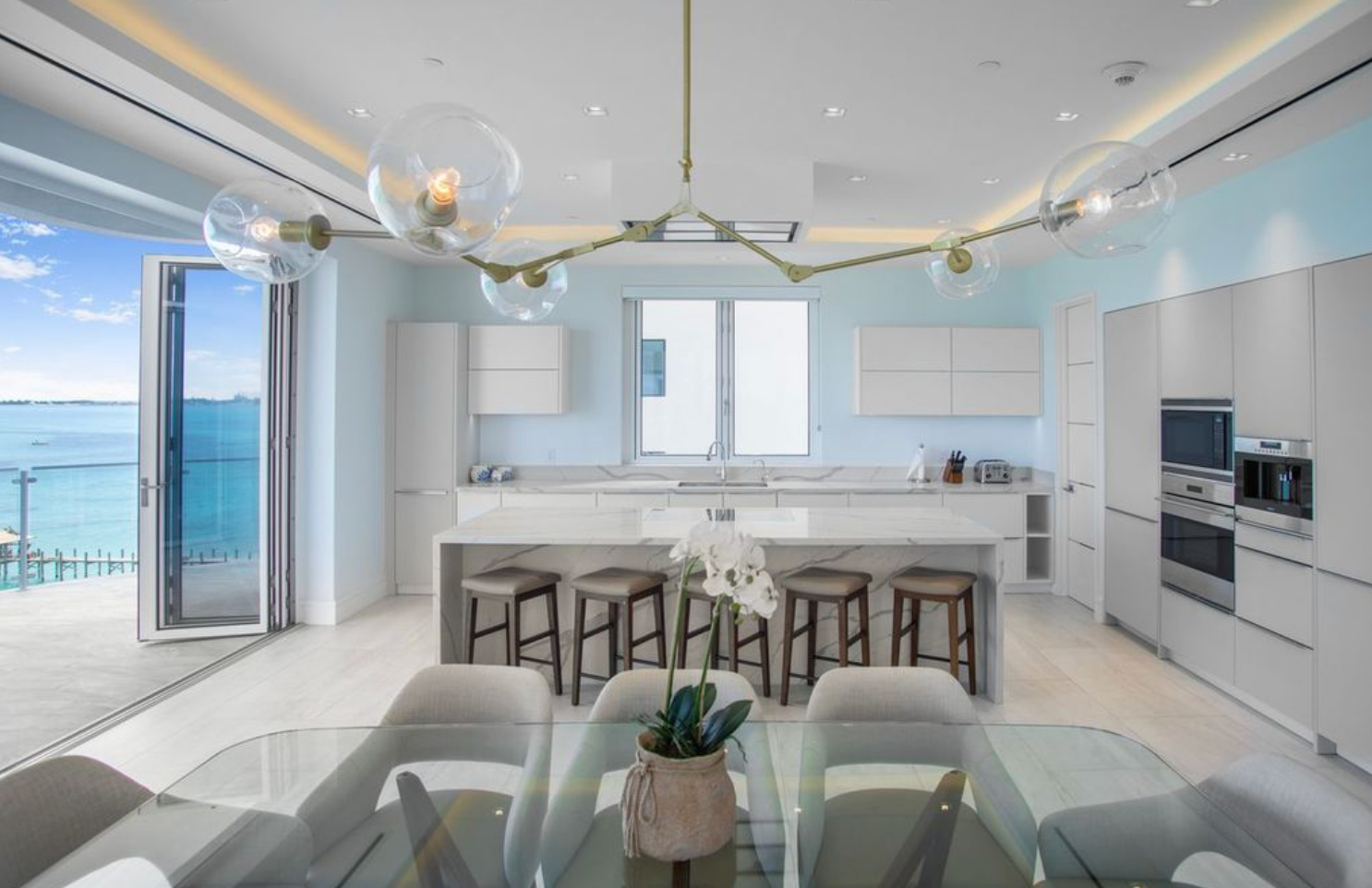 Modern kitchen with ocean view, light blue walls, white cabinets, island with stools, and glass dining table in The Bahamas