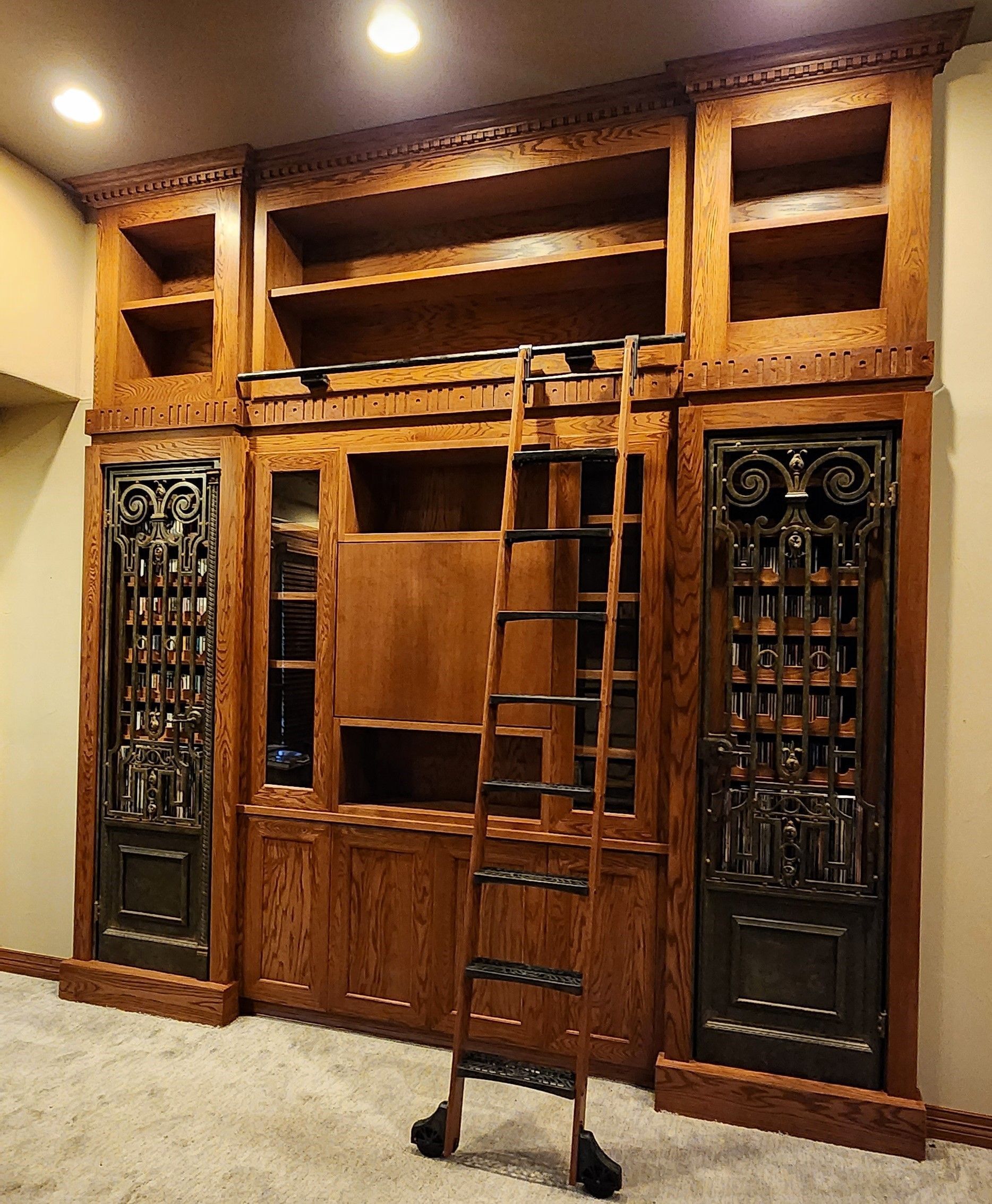 Wooden bookshelf unit with ladder, ornate doors, and built-in shelving, set against a beige wall.