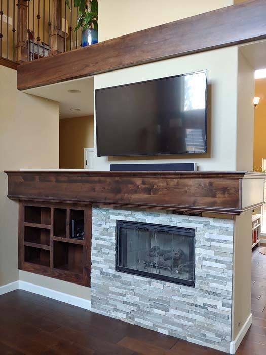 Living room fireplace with a TV above it, featuring stacked stone, a dark wood mantle, and shelving.