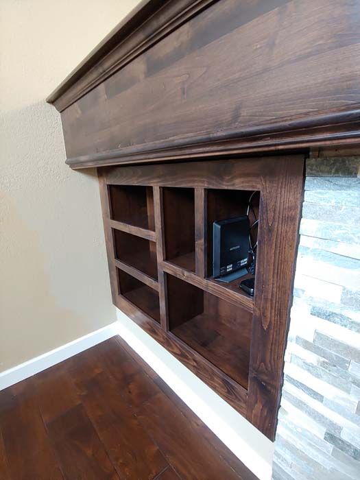 Built-in wooden shelf unit with a mantle above, stained dark brown, holding electronics. Located next to a stone wall.