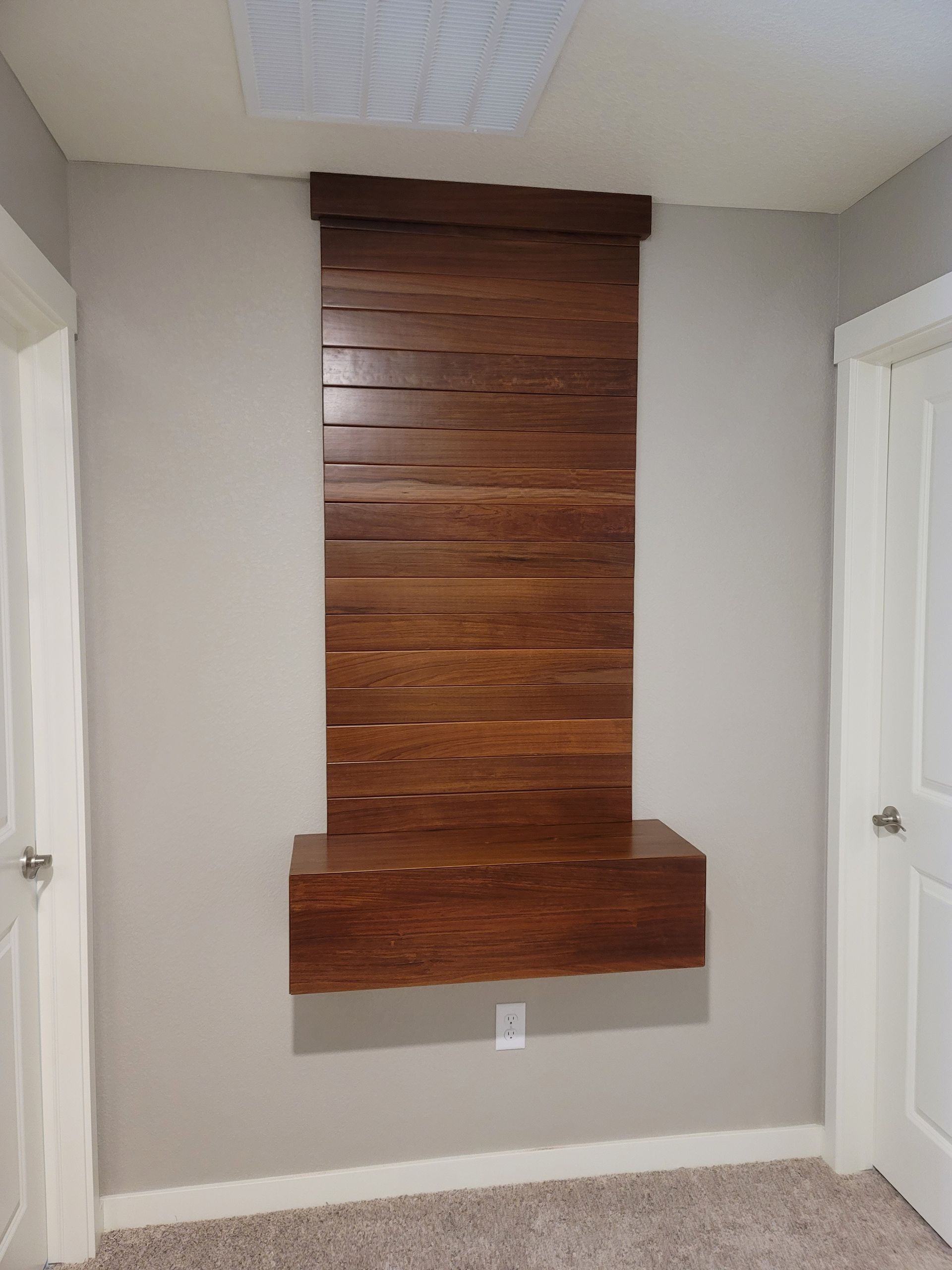 Wooden wall-mounted shelf and decorative panel in a room with light gray walls and carpet, between two white doors.