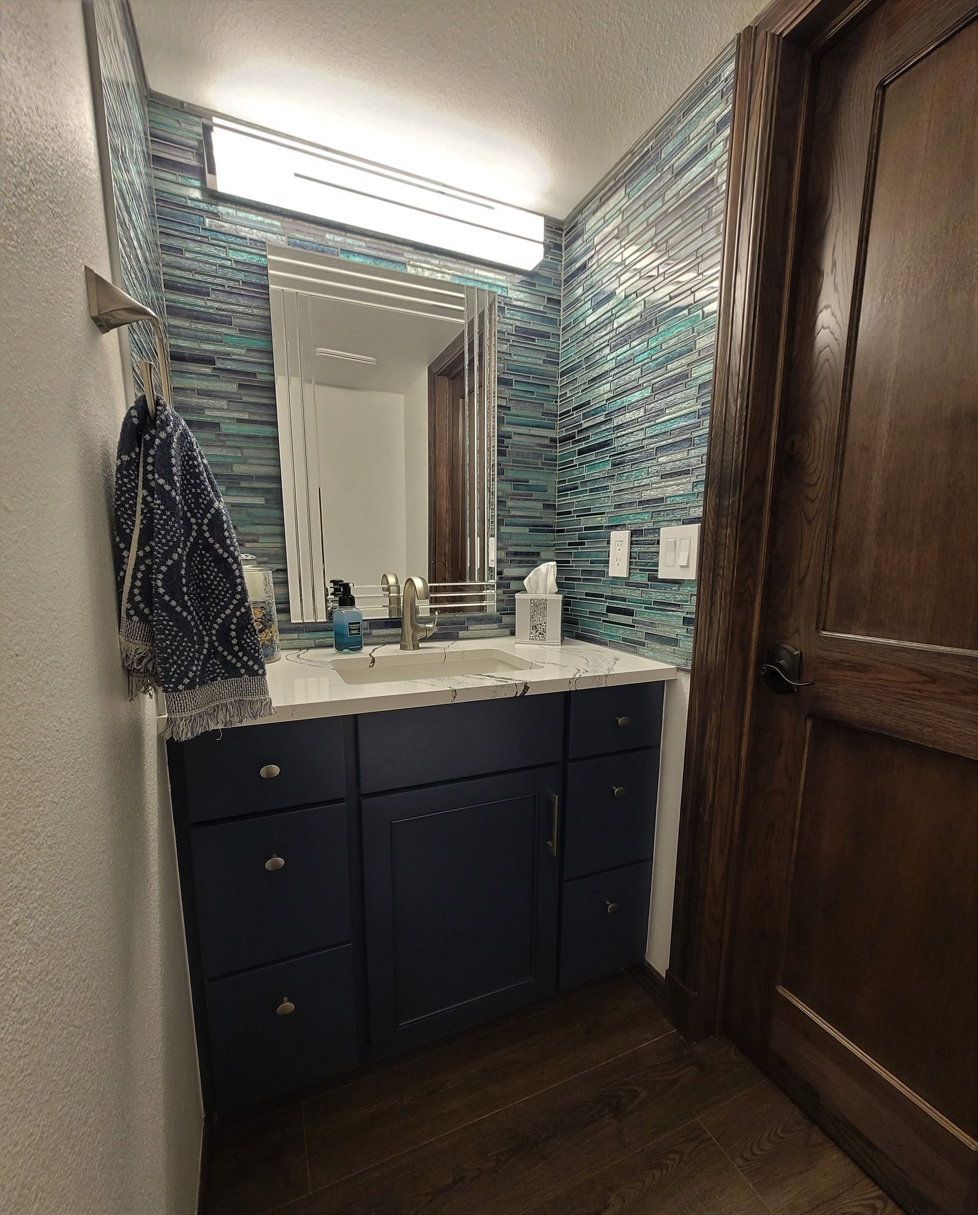 Bathroom with blue tiled backsplash, navy vanity, mirror, and a wooden door.