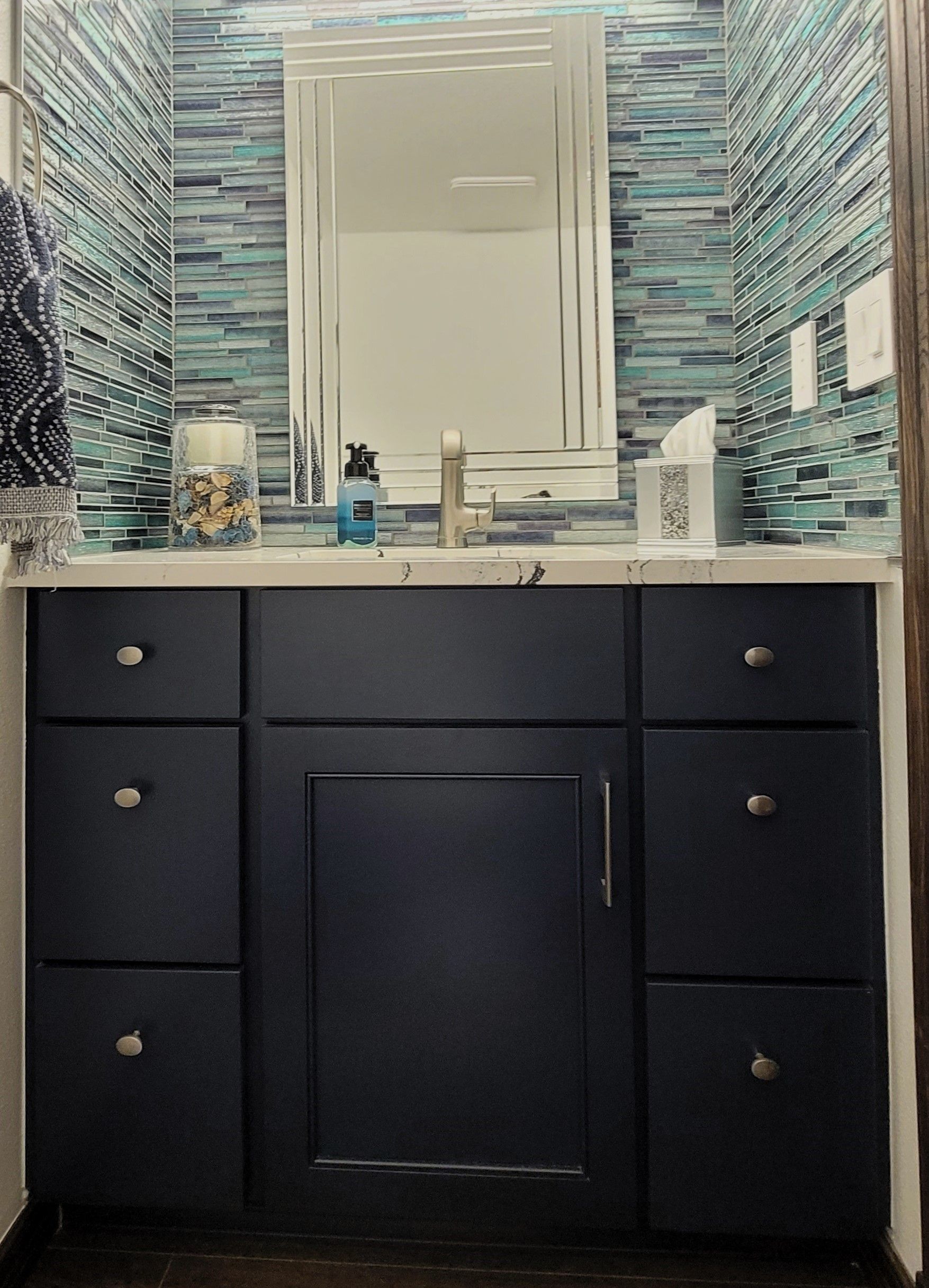 Navy vanity with silver knobs and a white countertop in a bathroom with blue tile backsplash and a hanging mirror.