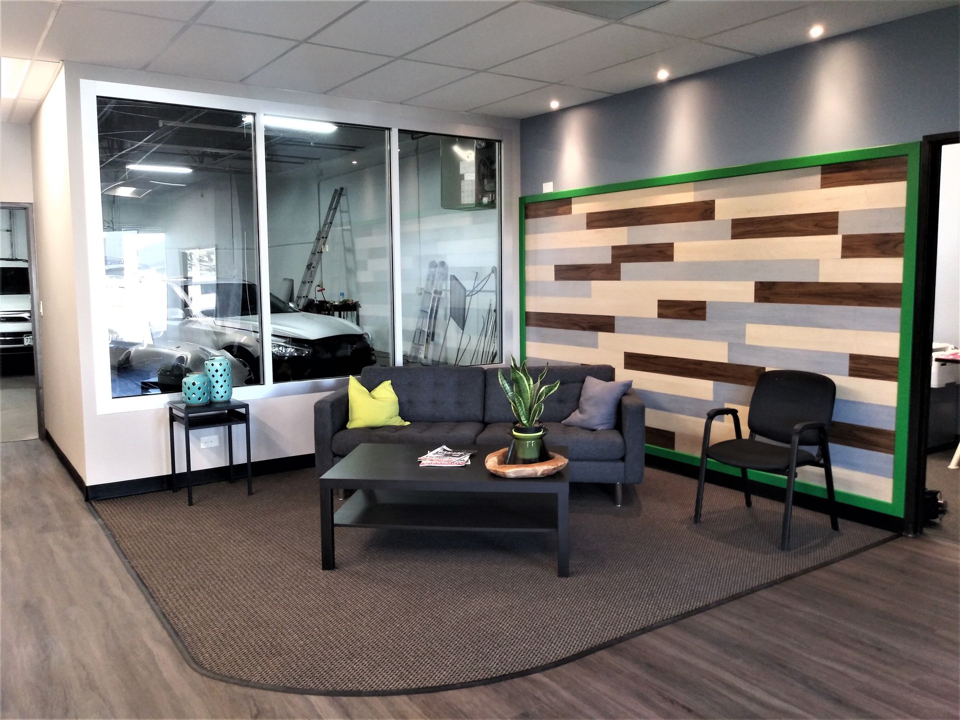 Waiting area with sofa, coffee table, chair, and decorative wall, with a view through a large window.