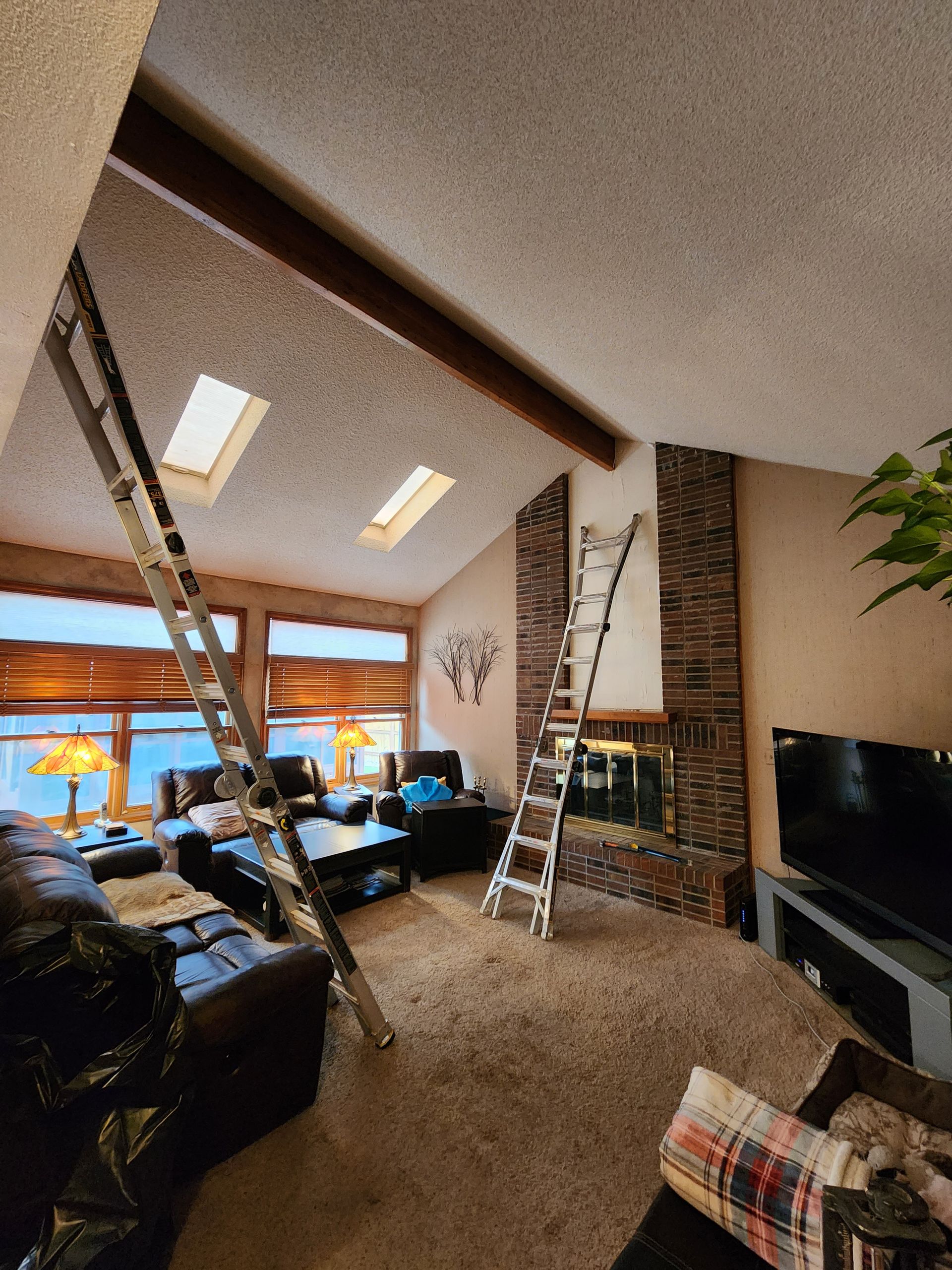 Two ladders propped against a slanted ceiling in a living room with a fireplace, skylights, and a television.
