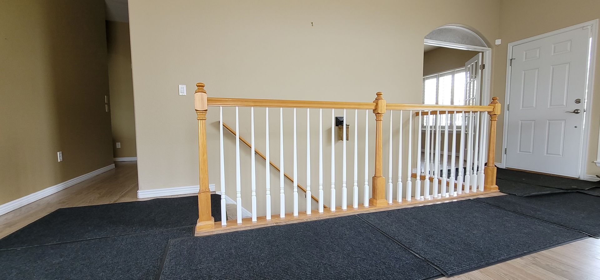 A light-colored wooden banister with white spindles sits in a house. A dark rug is in front.