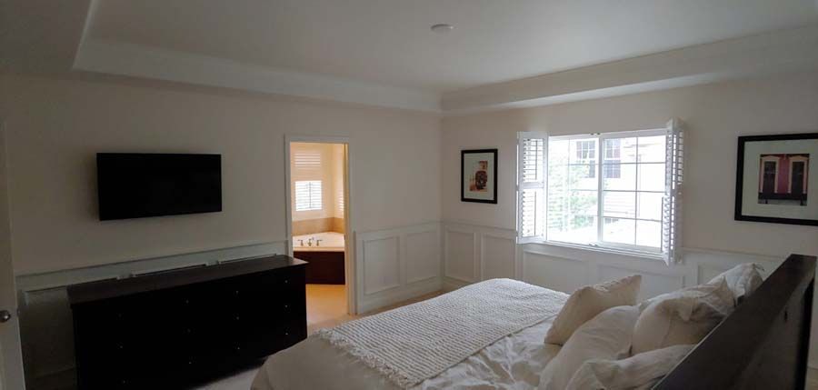 Bedroom with bed, television, and bathroom door.  White walls, window, and black dresser.
