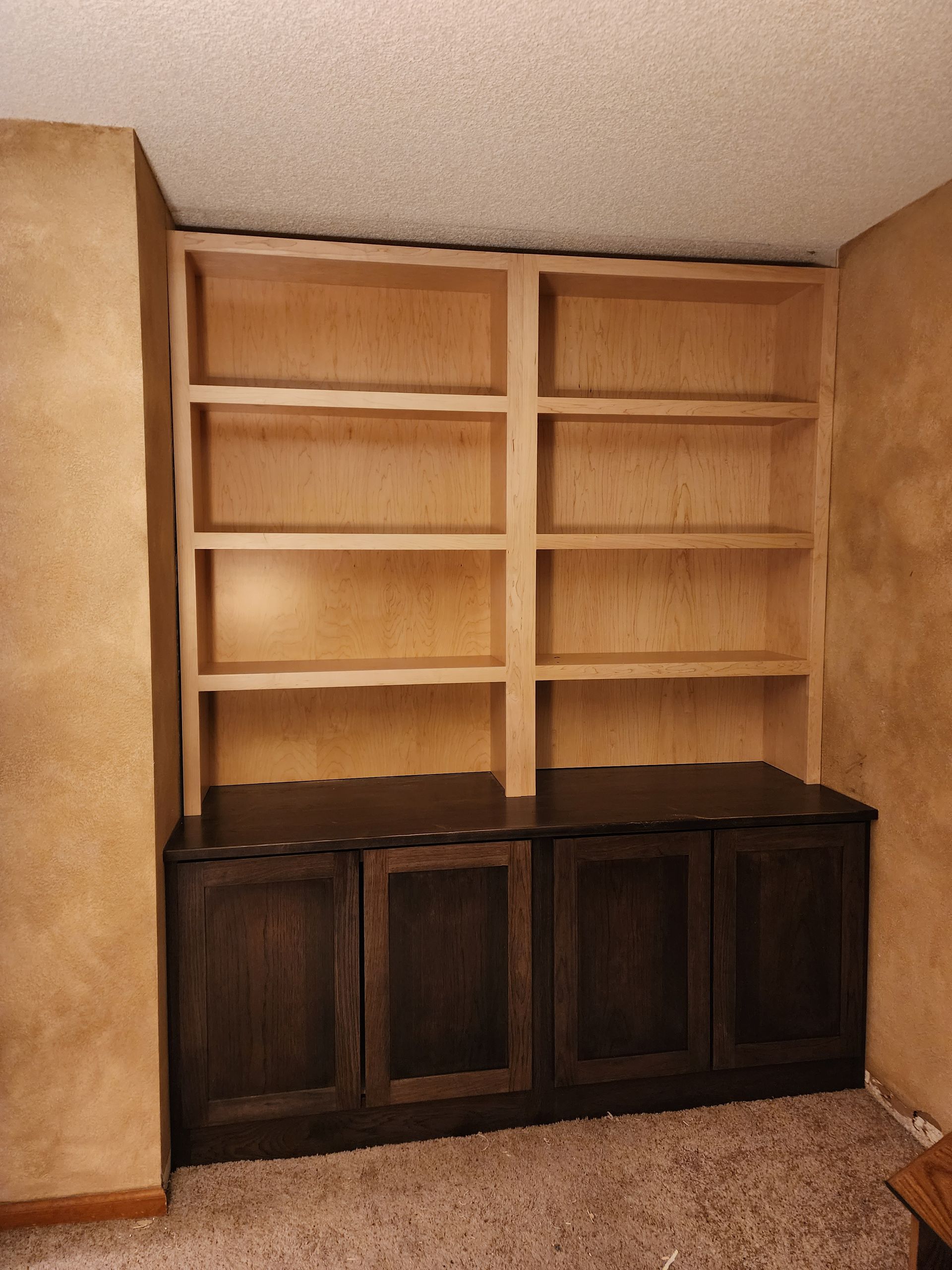 Built-in bookcase with light wood shelves and dark brown cabinet doors against textured tan walls.
