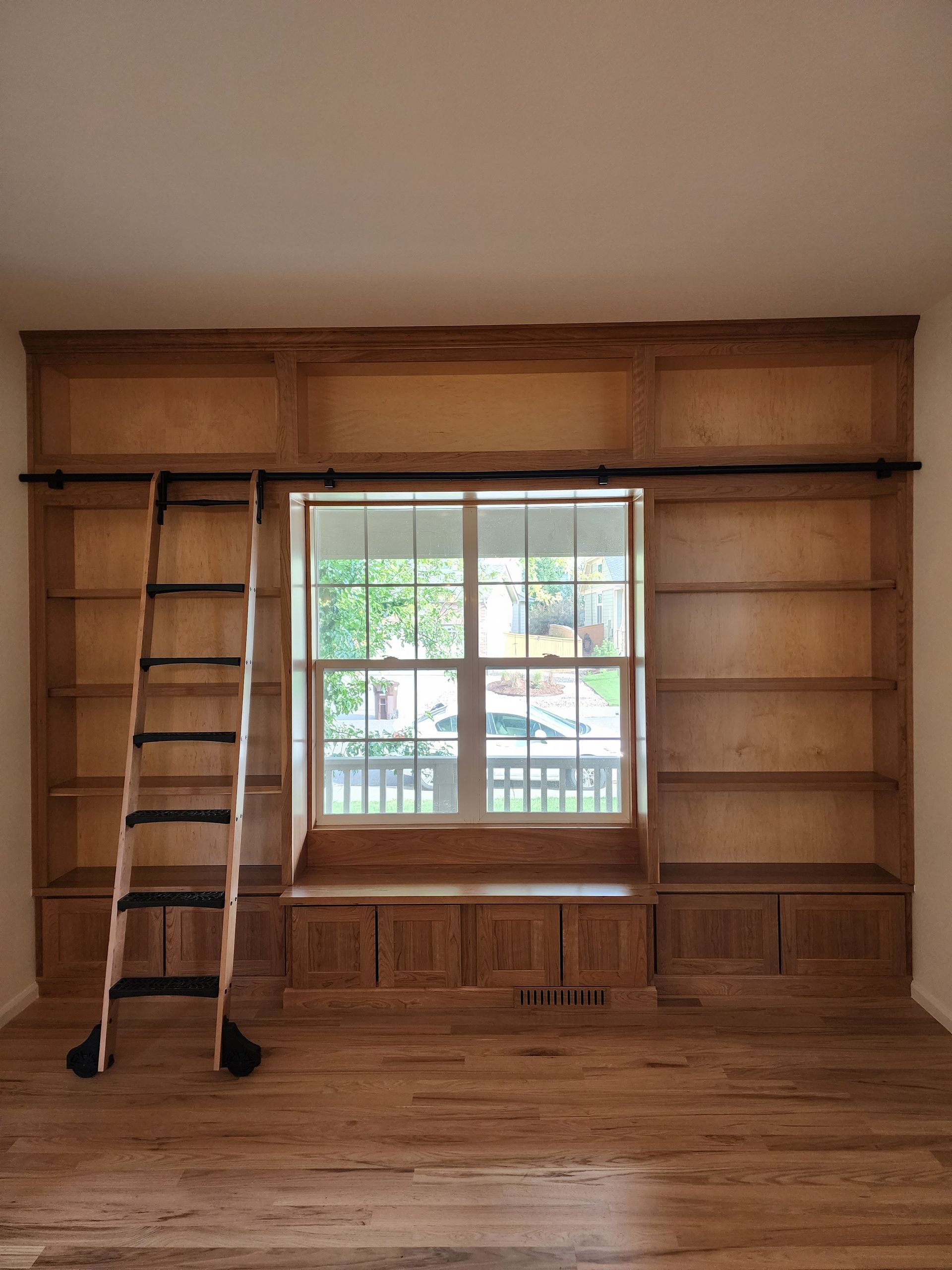 Built-in wooden bookcase with window seat and rolling ladder in a room with hardwood floors.