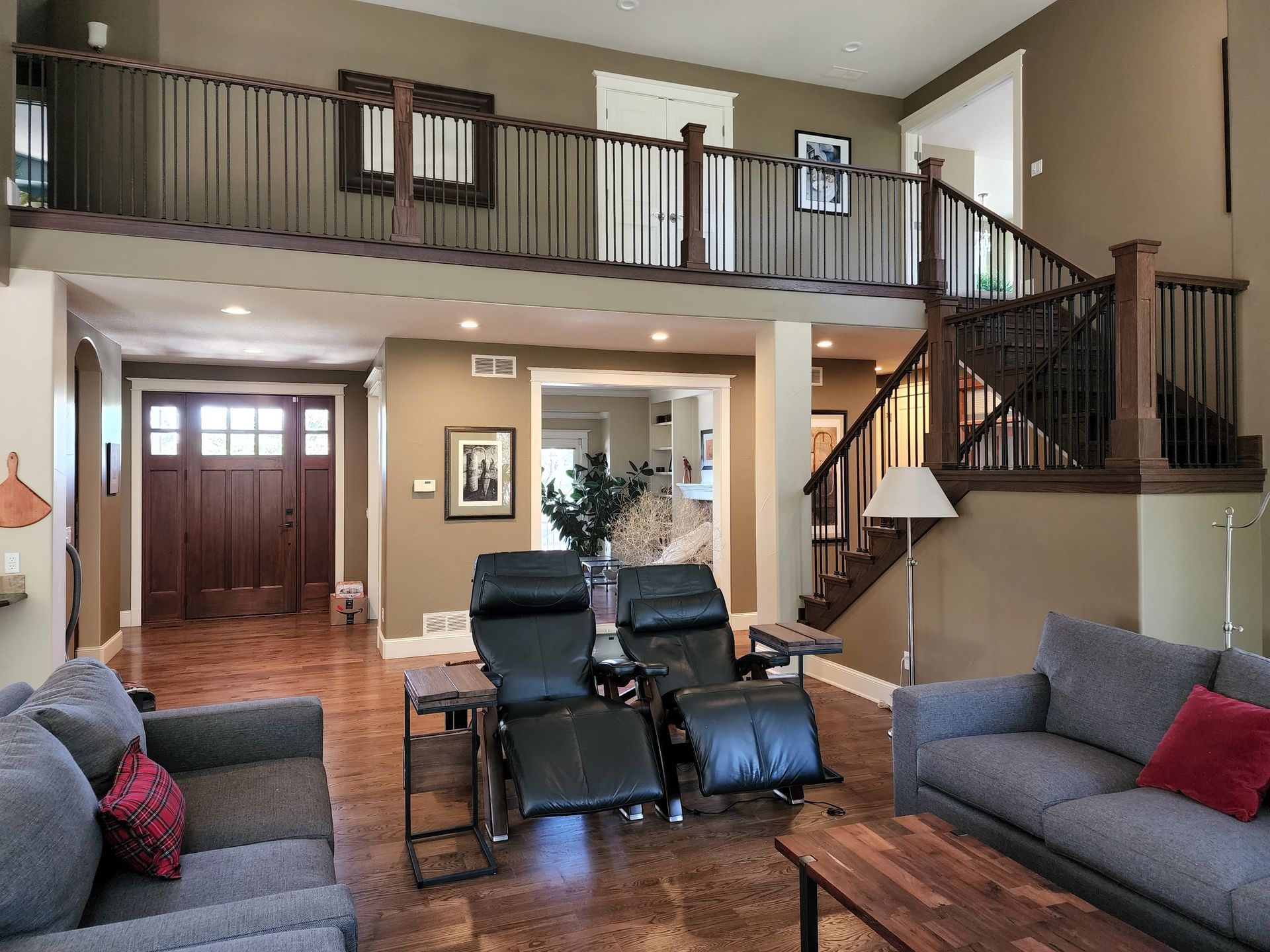 Spacious living room with a loft, staircase, and a wood door, featuring neutral walls, gray furniture, and recliners.