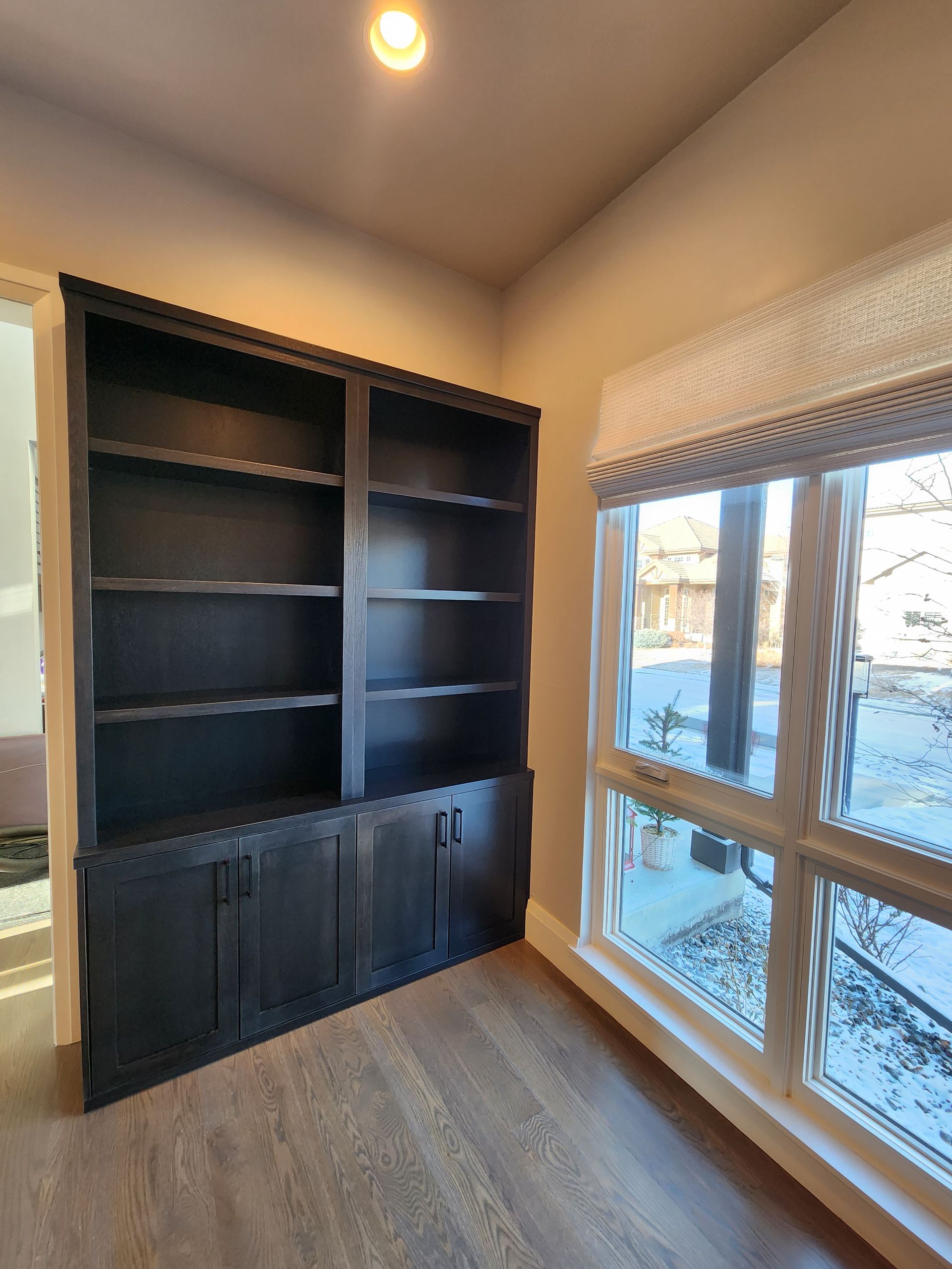 Dark wood built-in bookcase with shelves and cabinets, beside a window with a Roman shade.